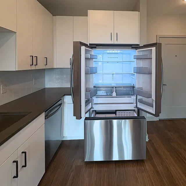 A modern kitchen featuring an open refrigerator with a stainless steel finish, showcasing its empty interior. The surrounding cabinetry is sleek and minimalistic, with a countertop and wooden flooring visible.