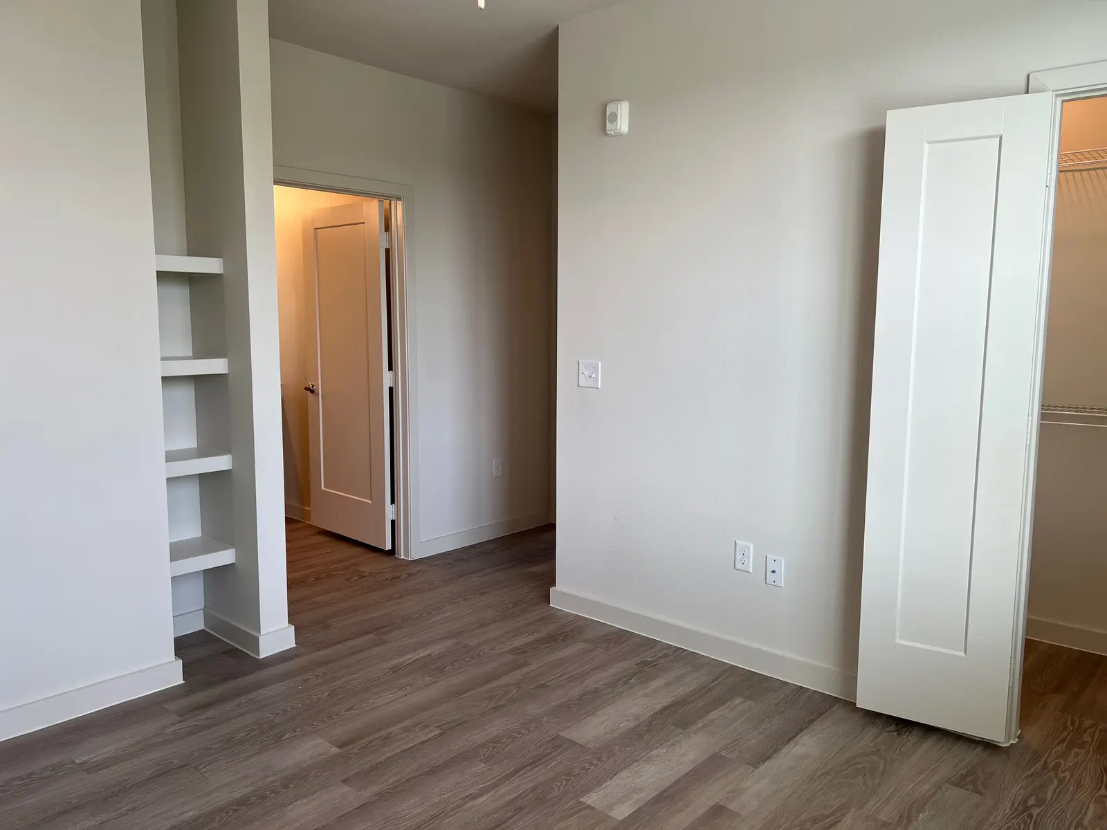 A spacious room interior featuring light-colored walls and hardwood flooring. There is an open door leading to another room and a built-in shelf on the left side. A closet door is visible on the right.
