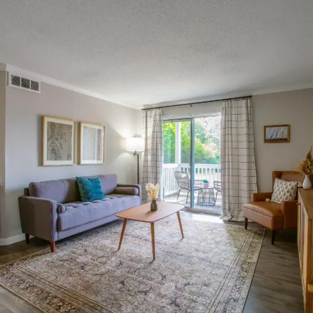 A modern living room featuring a grey couch, wooden coffee table, and a large window with a view of a balcony. The room is decorated with framed art, a light source beside the couch, and a patterned rug on the floor.