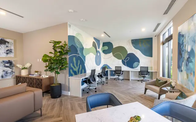 A modern office space featuring a mural with abstract shapes in green and blue hues, desks with black chairs, a small table with a plant, and additional seating areas with decorative plants.
