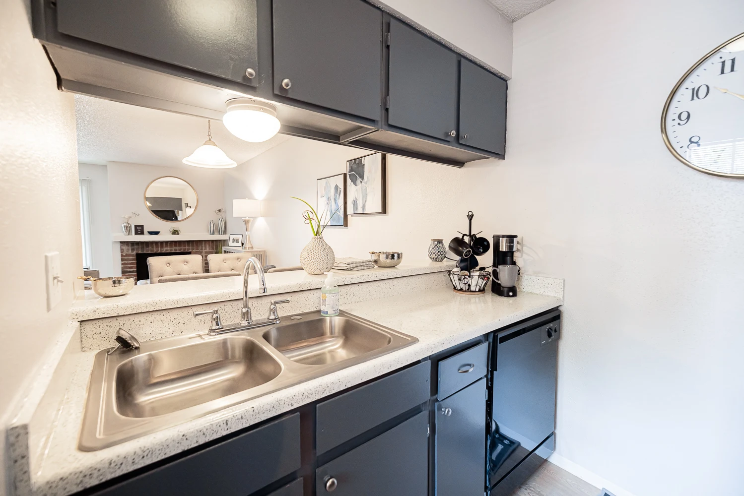 A modern kitchen featuring a double sink, dark cabinets, and white countertops. A coffee maker and utensils are also visible. There's a mirror reflecting a living area in the background.