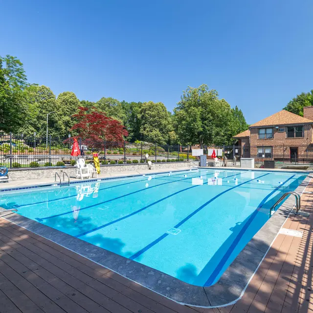 A clear blue swimming pool surrounded by a wooden deck and lawn, with a few chairs and lounge areas. Trees and a building are visible in the background.