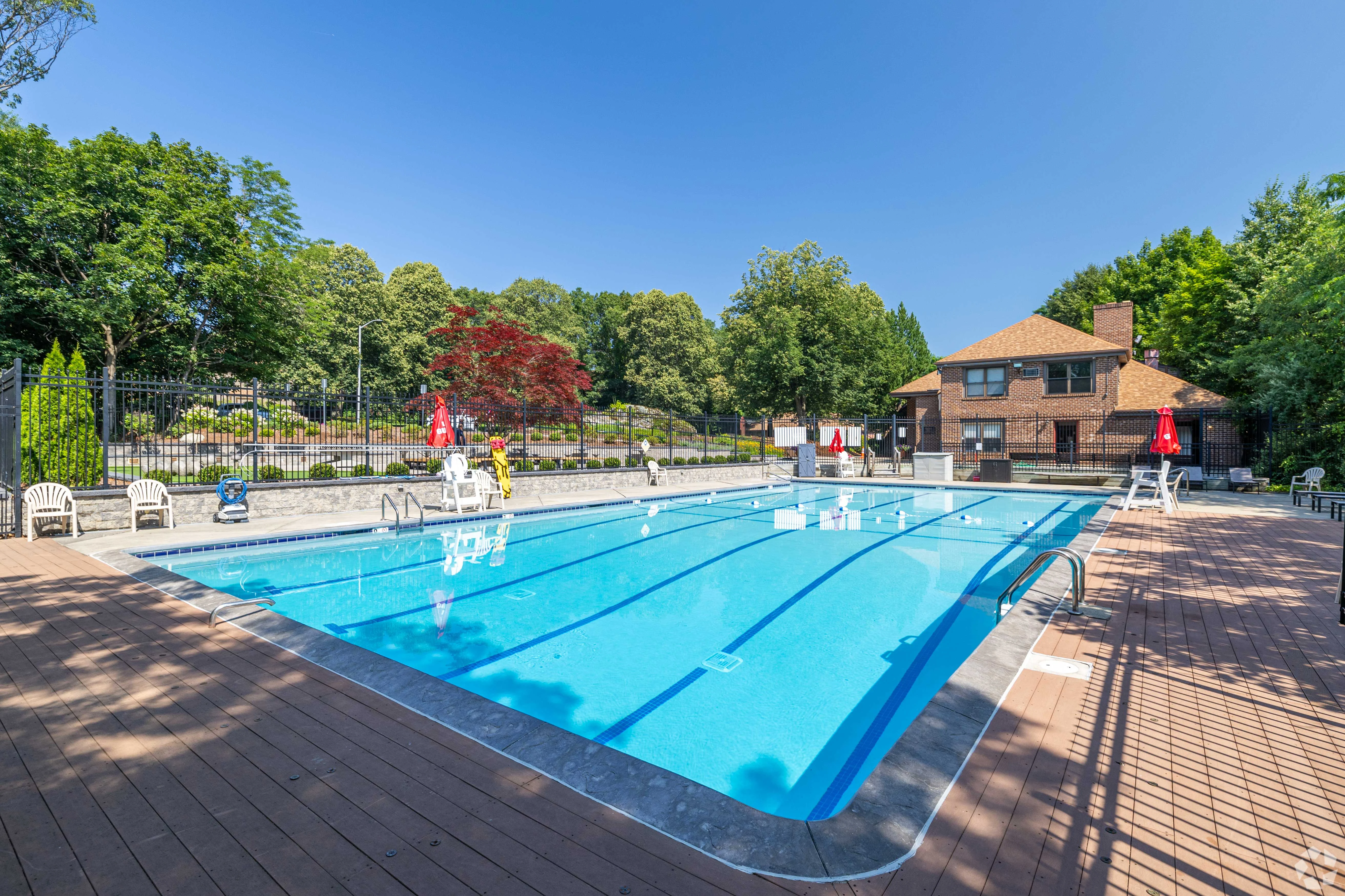 Sunny Swimming Pool Area A clear blue swimming pool surrounded by a wooden deck and lawn, with a few chairs and lounge areas. Trees and a building are visible in the background.