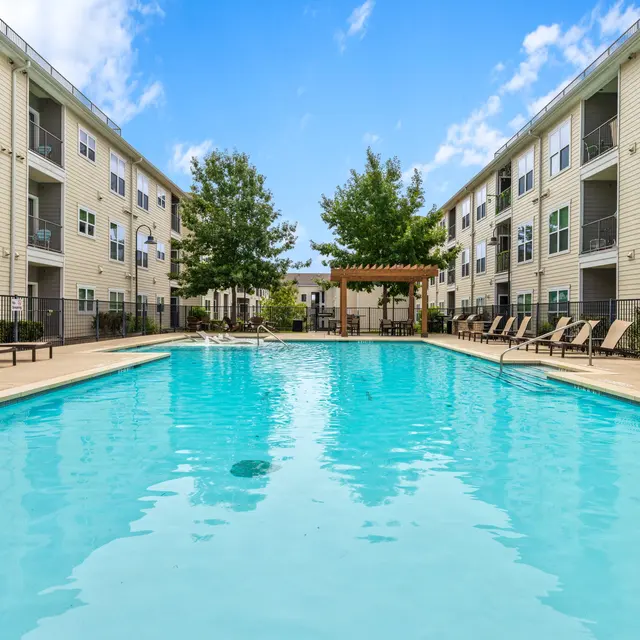 A view of a pool surrounded by beige apartment buildings, with a pergola and several lounge chairs