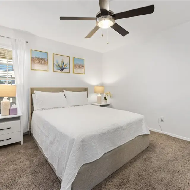 A cozy bedroom featuring a queen-sized bed with white bedding, a nightstand with a lamp, and framed artwork on the wall. Large windows let in natural light, with light curtains. A fan is mounted on the ceiling.