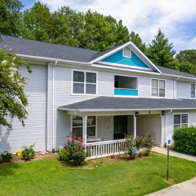 Two-story modern apartment building with a light grey exterior and blue accents, surrounded by greenery.