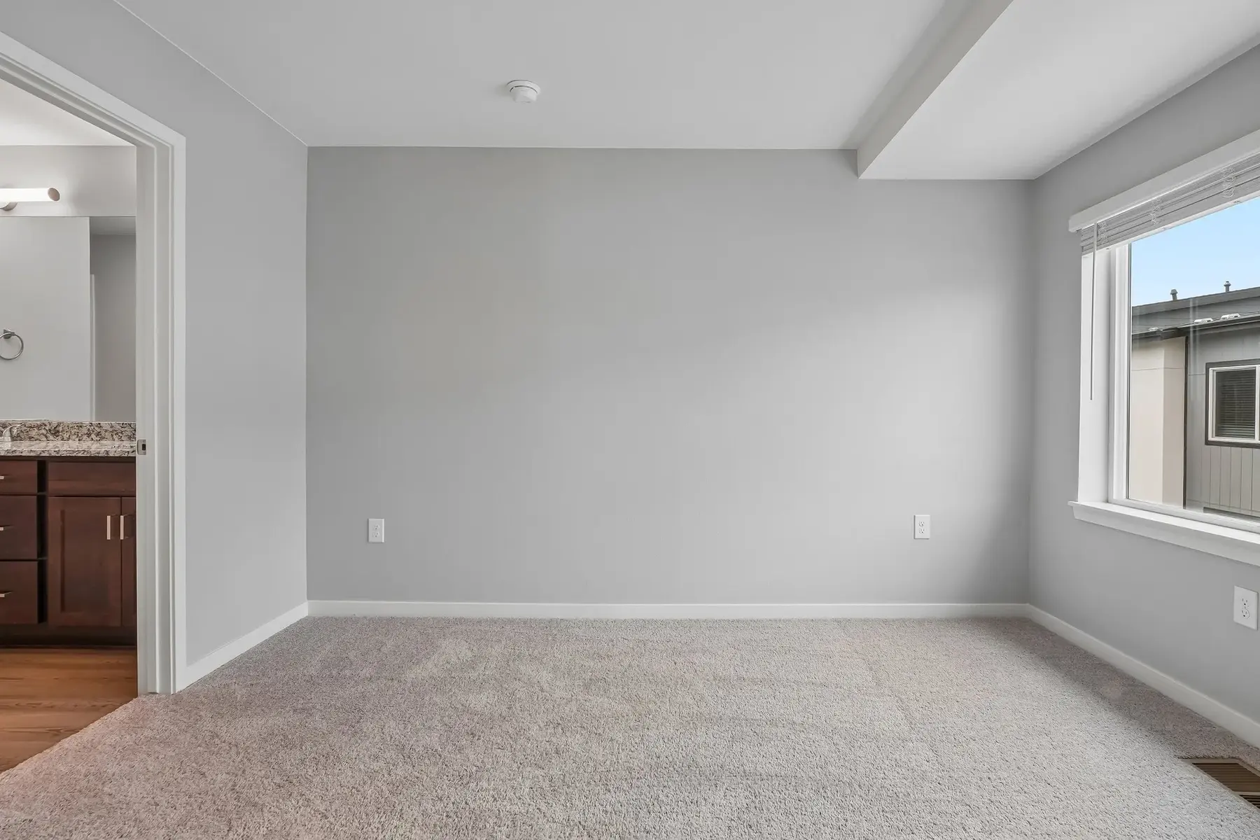 Spacious Empty Room An empty room with light gray walls, carpeted floor, and a window letting in natural light. A doorway on the left leads to another area, possibly a bathroom or closet.