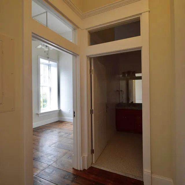 Interior view showing a hallway with an entry to a bathroom on the right and a room visible through an arched doorway on the left, featuring wooden flooring and large windows.