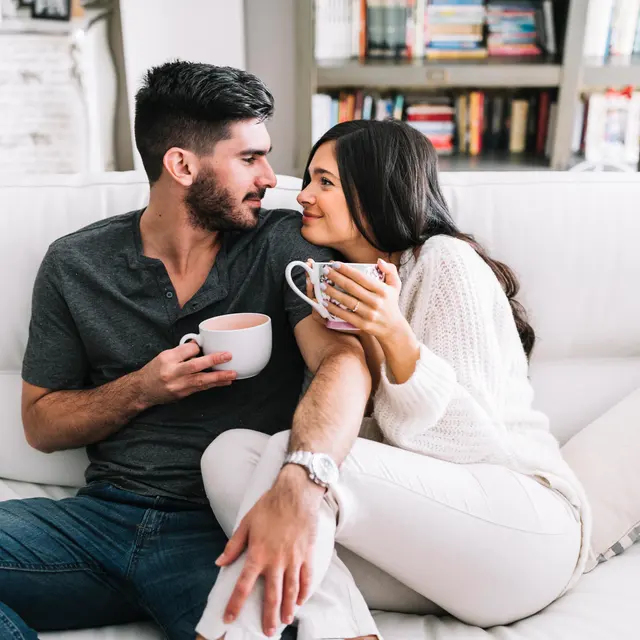 The Hideaway A young couple sitting on a white couch, enjoying cups of coffee together in a cozy living room atmosphere.
