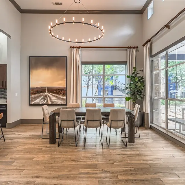 A spacious modern dining area with a large rectangular table surrounded by chairs, a contemporary chandelier above, and large windows letting in natural light. Artwork is displayed on the wall and a plant is positioned near the window.