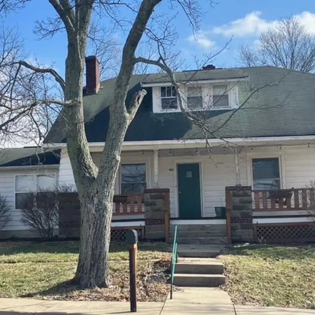 A two-story house with a green roof, front porch, and large tree in the yard. The house has a simple design with a white exterior and a green door.