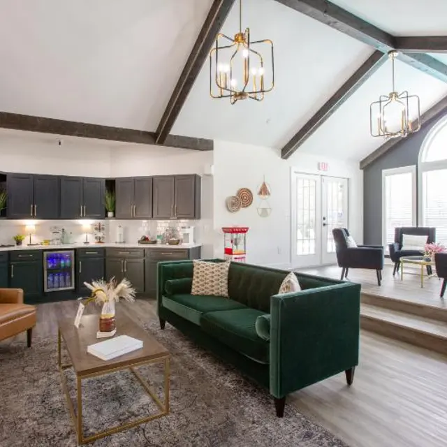 A modern living space featuring a green velvet sofa, beige armchair, and stylish decor. The room has high ceilings with wooden beams and large windows letting in natural light. A kitchen area is visible with dark cabinetry and under-cabinet lighting.