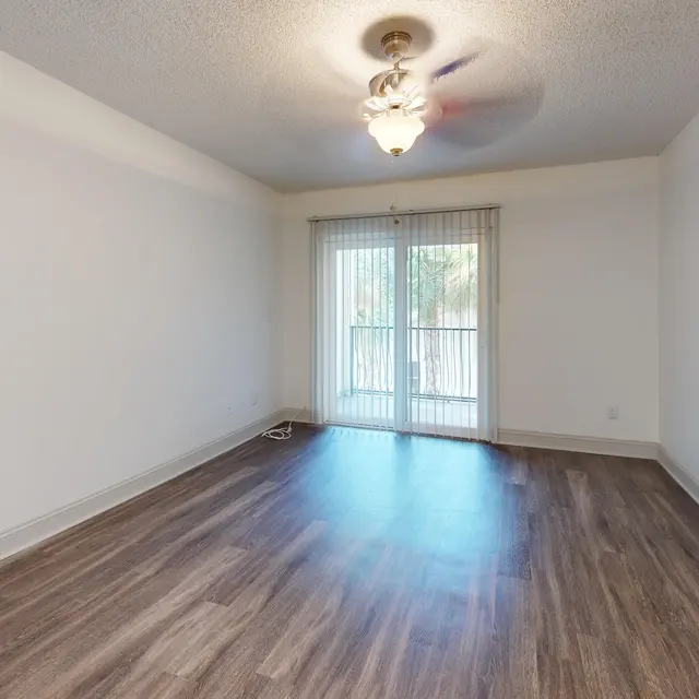 A spacious empty living room with wood-like flooring and a ceiling fan. There are large sliding doors leading to an outside balcony, allowing natural light to enter the room. The walls are painted white, creating a bright and airy atmosphere.