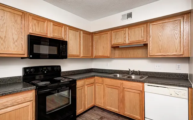 A modern kitchen featuring wooden cabinets, a black stove and microwave, and a dishwasher. The countertop is dark and the floor is a light wood-like material.