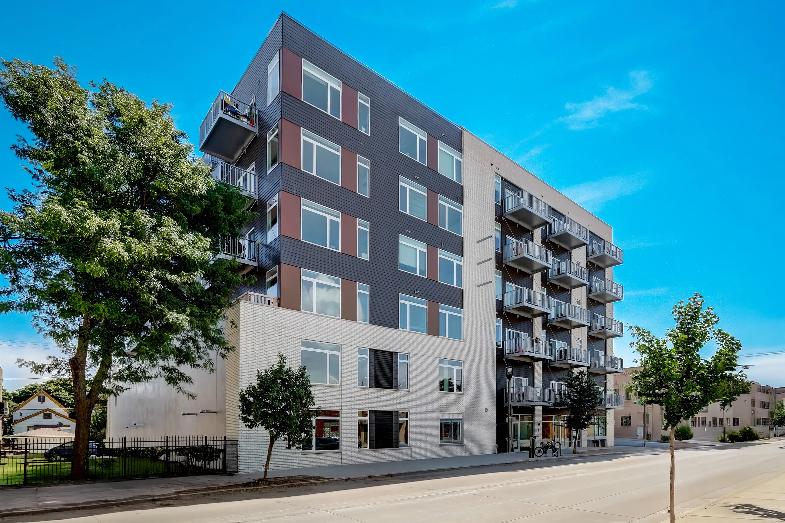 A modern apartment building with balconies and large windows, surrounded by trees on a sunny day.