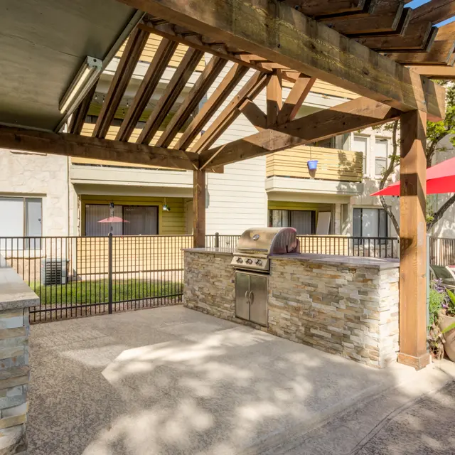 A well-designed outdoor grilling area featuring a stone grill station under a wooden pergola, with a seating area and patio umbrellas in the background.