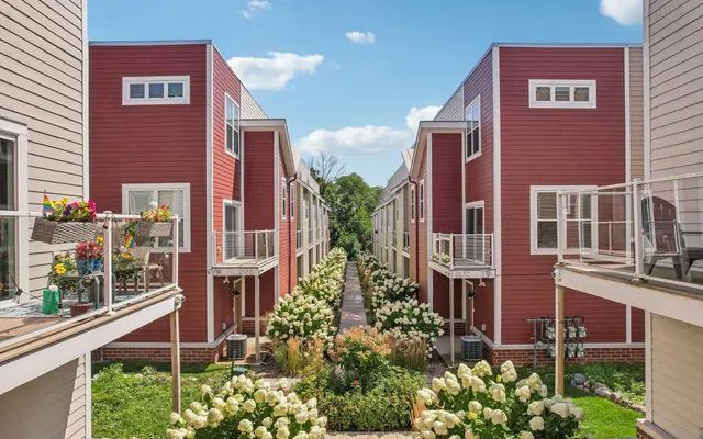 A vibrant modern housing complex with two lines of buildings flanking a landscaped path filled with flowers, under a blue sky with scattered clouds.