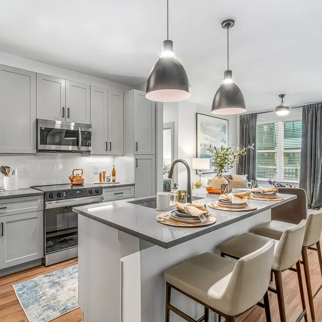 A modern kitchen featuring gray cabinetry, stainless steel appliances, and a large island with bar stools. Decor includes a vase of flowers and a rug on the floor.