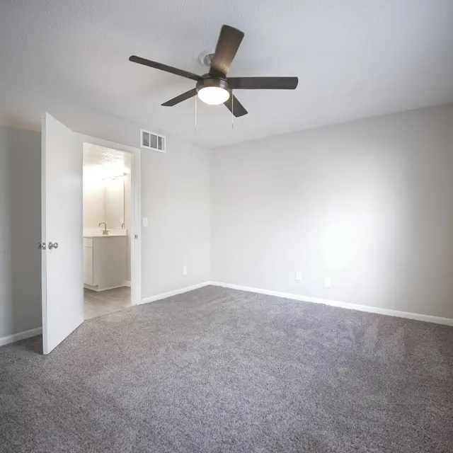 A spacious empty bedroom with gray carpet, a ceiling fan, and a door leading to a bathroom. The walls are painted light gray and there are windows with blinds letting in natural light.