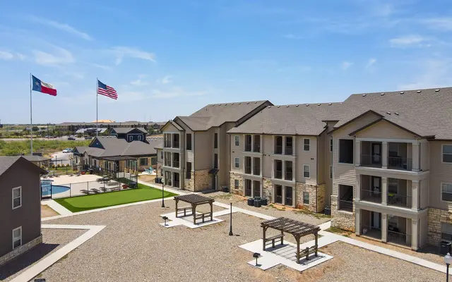 Aerial view of a modern apartment complex with two flags flying, a pool area, and landscaped outdoor space.