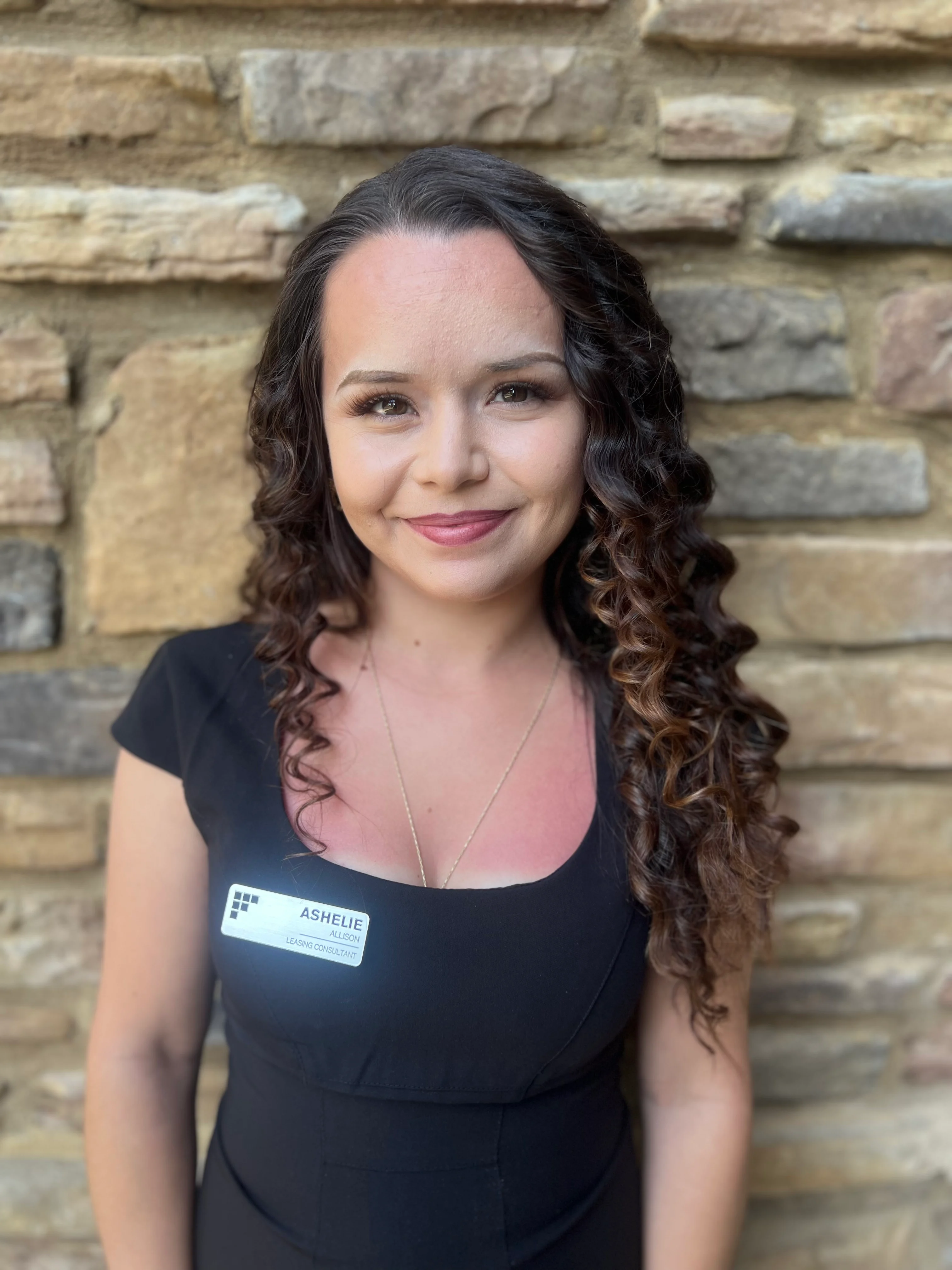 A woman with long curly hair, wearing a black dress and a name tag, stands in front of a stone wall. She has a friendly smile and a relaxed posture.