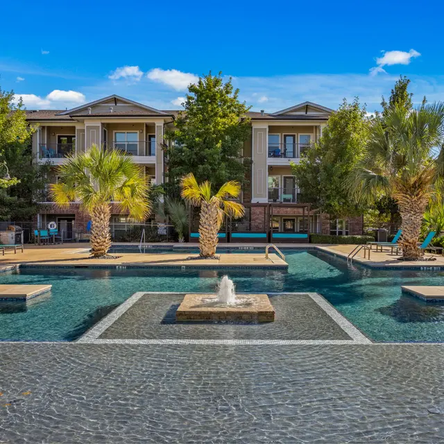 A spacious apartment complex pool area featuring palm trees, a fountain, and lounge chairs under a clear blue sky.
