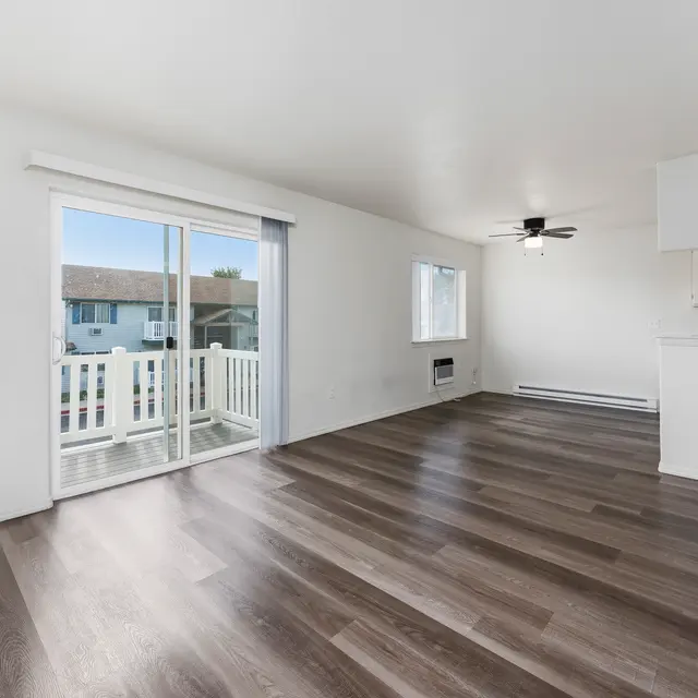 Bright and Spacious Living Room A bright and spacious living room with large windows, light wooden flooring, and a ceiling fan. It features a sliding door that opens to a balcony and has an open layout leading to a small kitchen area.