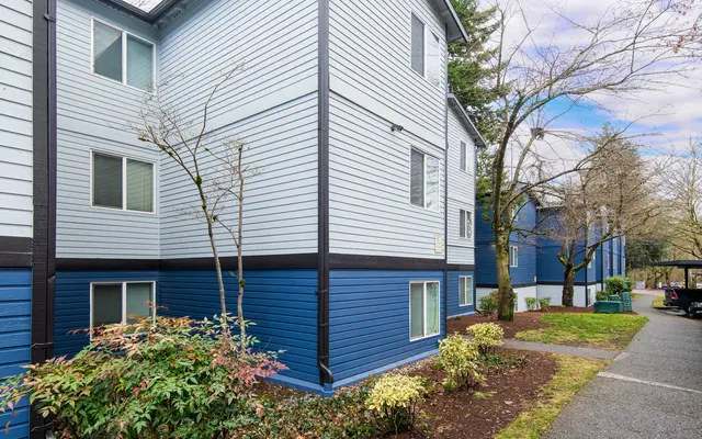 Exterior view of a blue and light blue apartment building with windows and greenery in the foreground.