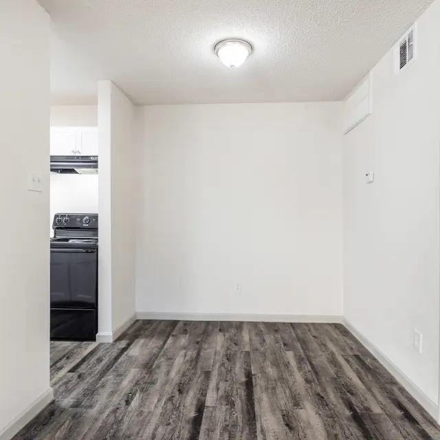 A view of a simple, empty apartment interior showing a living area with new flooring. The wall is painted white and there is a light fixture on the ceiling. In the background, a kitchen area with a stove is visible, and a bathroom is seen at the right of the image.