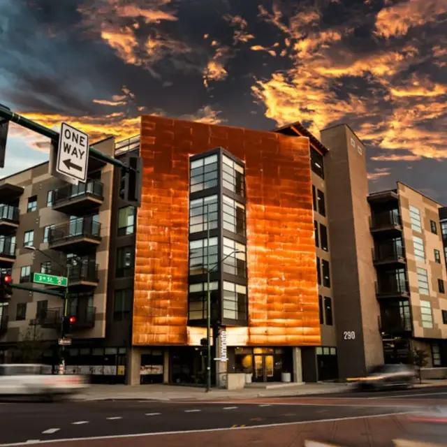 A modern building with a copper-colored facade reflecting a vibrant sunset, surrounded by other structures and street signs.