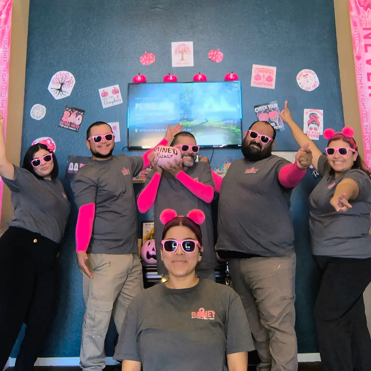 A group of six individuals posing in a playful manner, wearing matching gray shirts and pink accessories, with decorations in the background that include motivational messages and a television screen.