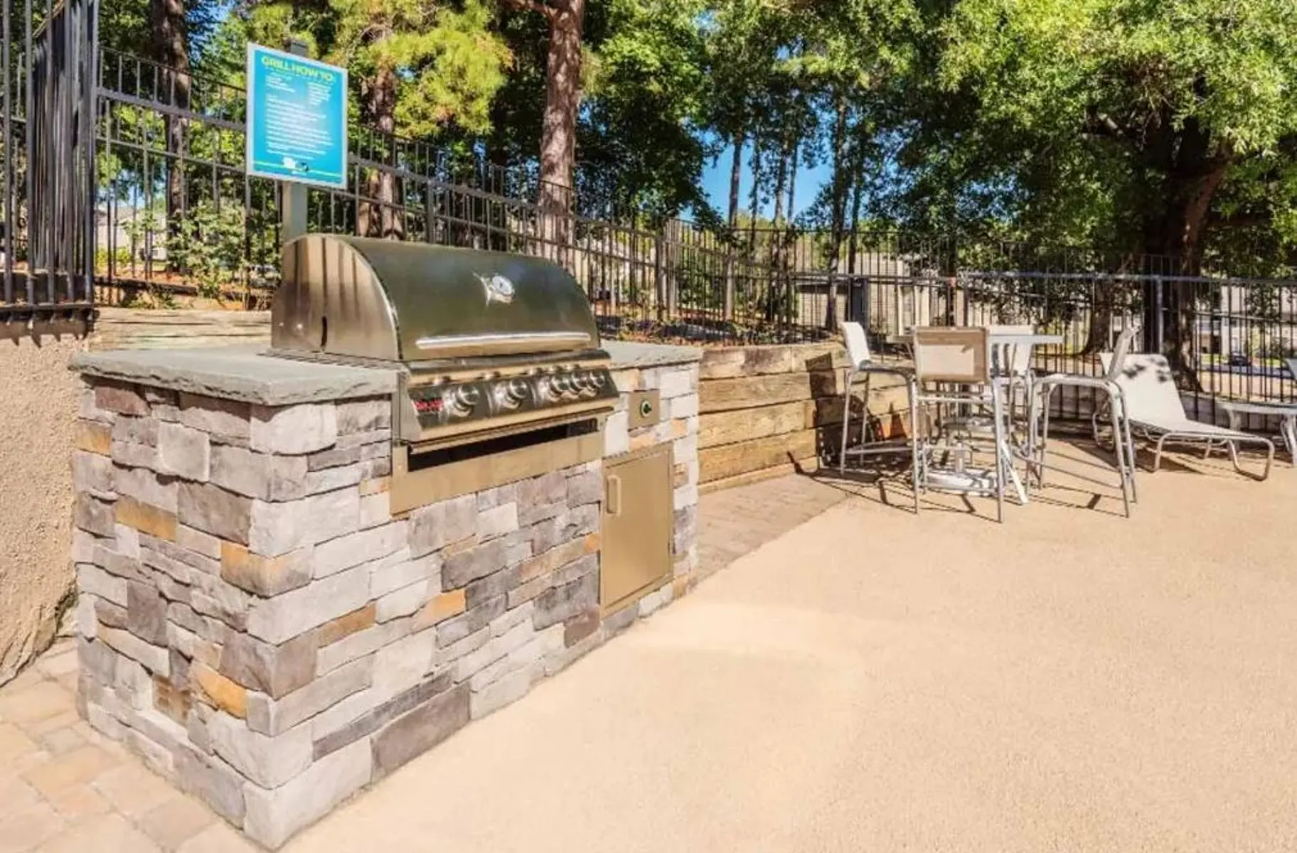 An outdoor grill area featuring a stone-clad barbecue grill, adjacent seating, and surrounded by greenery.
