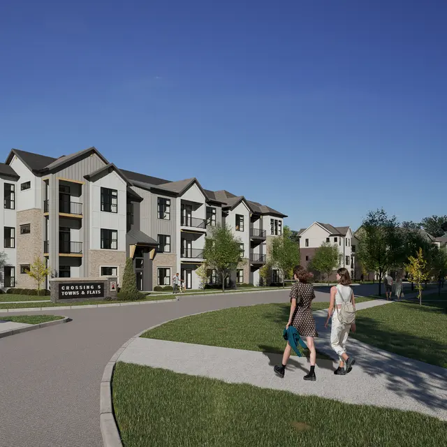 A modern apartment complex with landscaped grounds, featuring multiple buildings in a suburban setting under a clear blue sky. People are walking along the pathways.