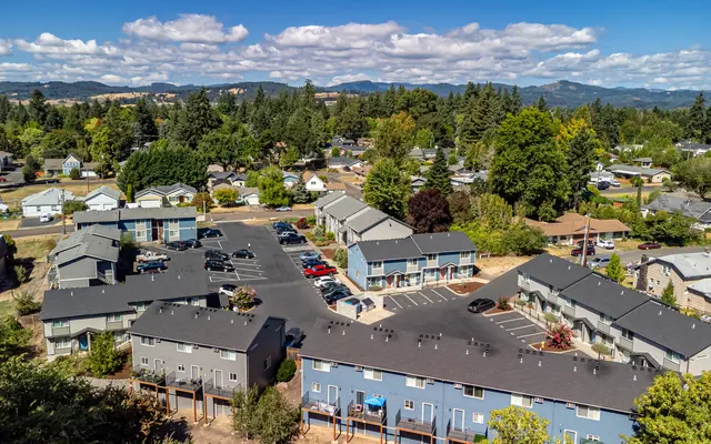 Aerial view of a residential area featuring an apartment complex surrounded by trees and smaller houses under a blue sky with clouds.