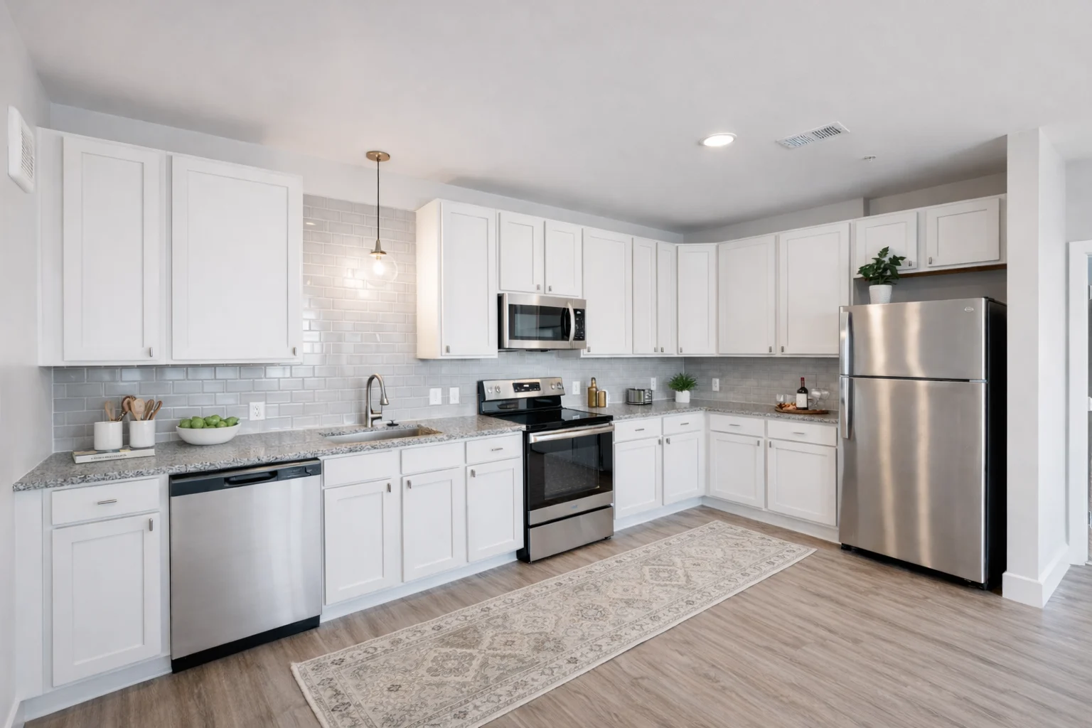 A modern kitchen featuring white cabinets, stainless steel appliances, and a light-colored countertop. The kitchen has a large rug on the floor and soft lighting from a pendant lamp.