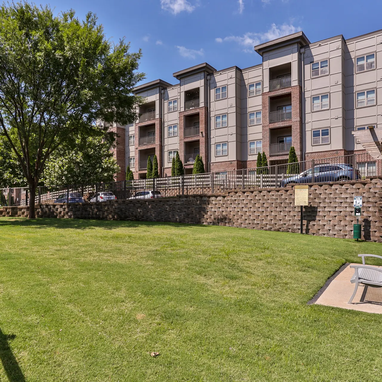 A well-maintained green space in front of an apartment building, featuring a lawn, trees, and fenced off areas.