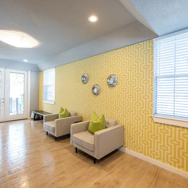 Modern Living Area A modern living area featuring two light gray armchairs with bright green cushions. The wall behind has a bold yellow geometric pattern with decorative wall pieces. A large window with blinds allows natural light to enter, and there's a door leading outside. The flooring is light wood.
