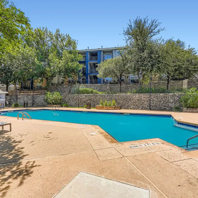 A sunny pool area surrounded by trees and an apartment building. The pool has clear water, a diving board, and lounging chairs.