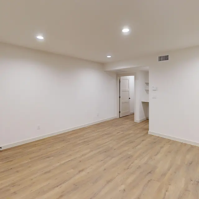 A spacious, modern apartment interior featuring light wood flooring, white walls, and recessed lighting.