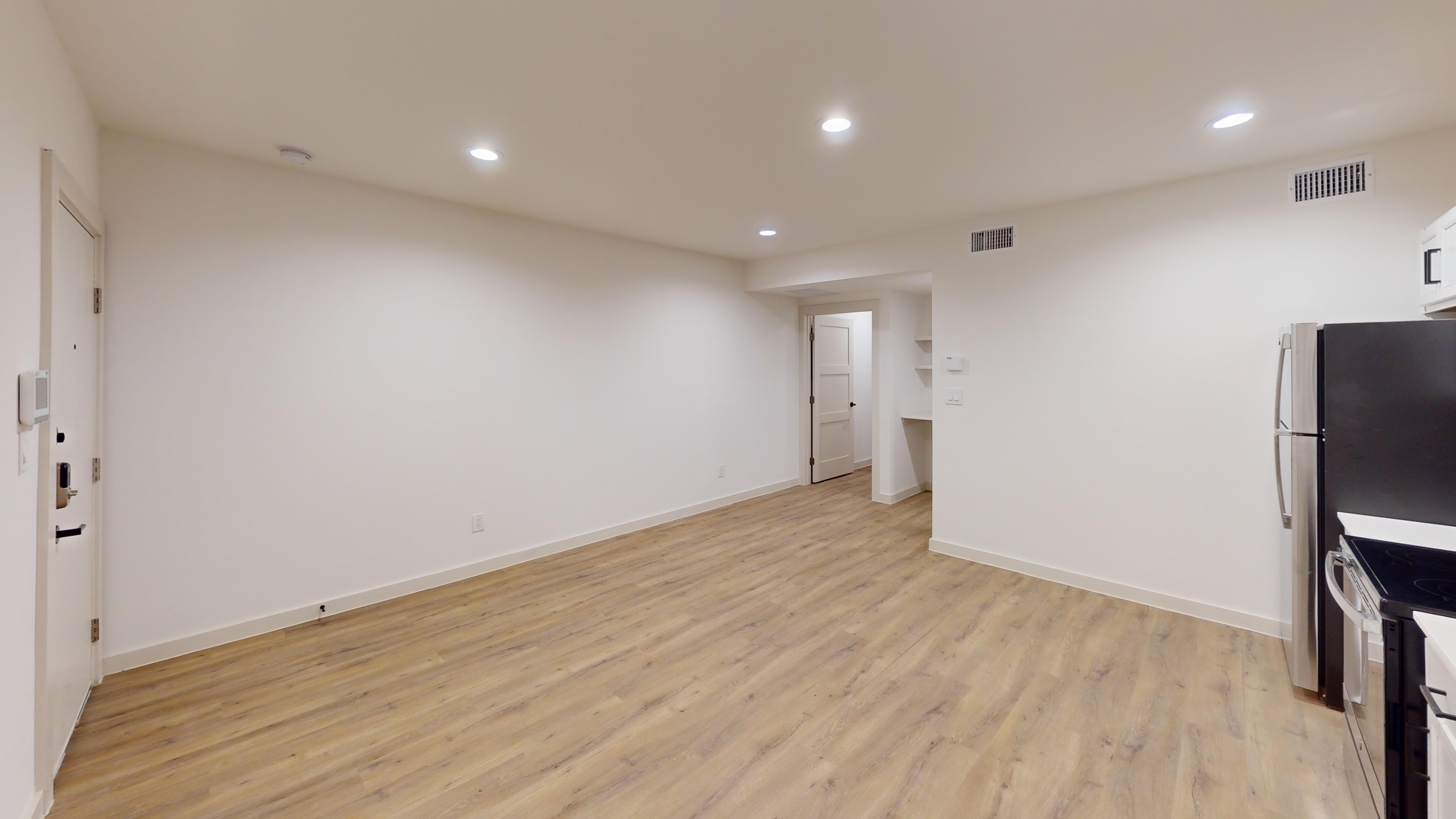 A spacious, modern apartment interior featuring light wood flooring, white walls, and recessed lighting.