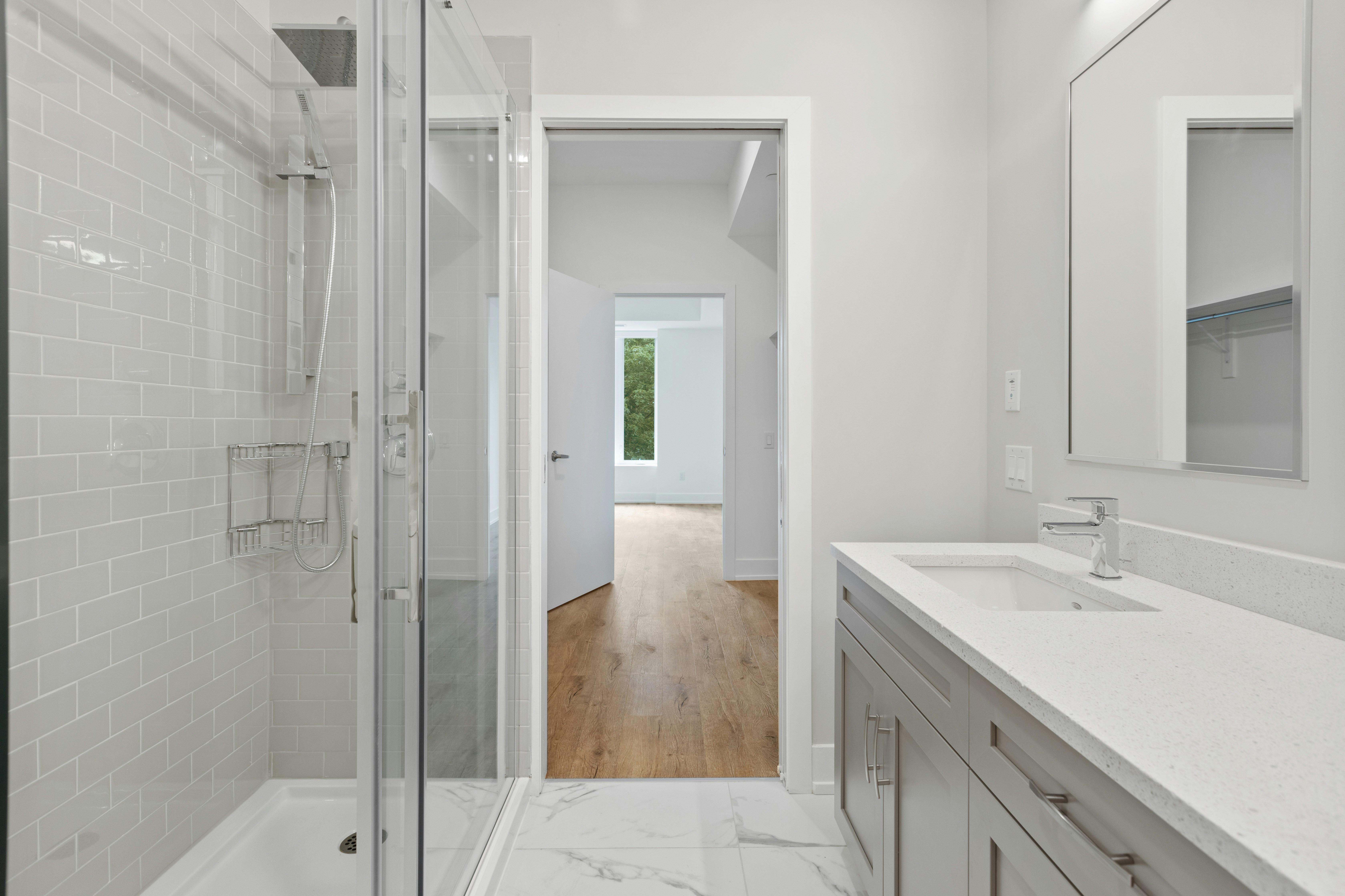 A modern bathroom featuring a glass shower enclosure, sleek countertop sink, and light-toned cabinetry. The background shows a hallway leading to a brightly lit room.