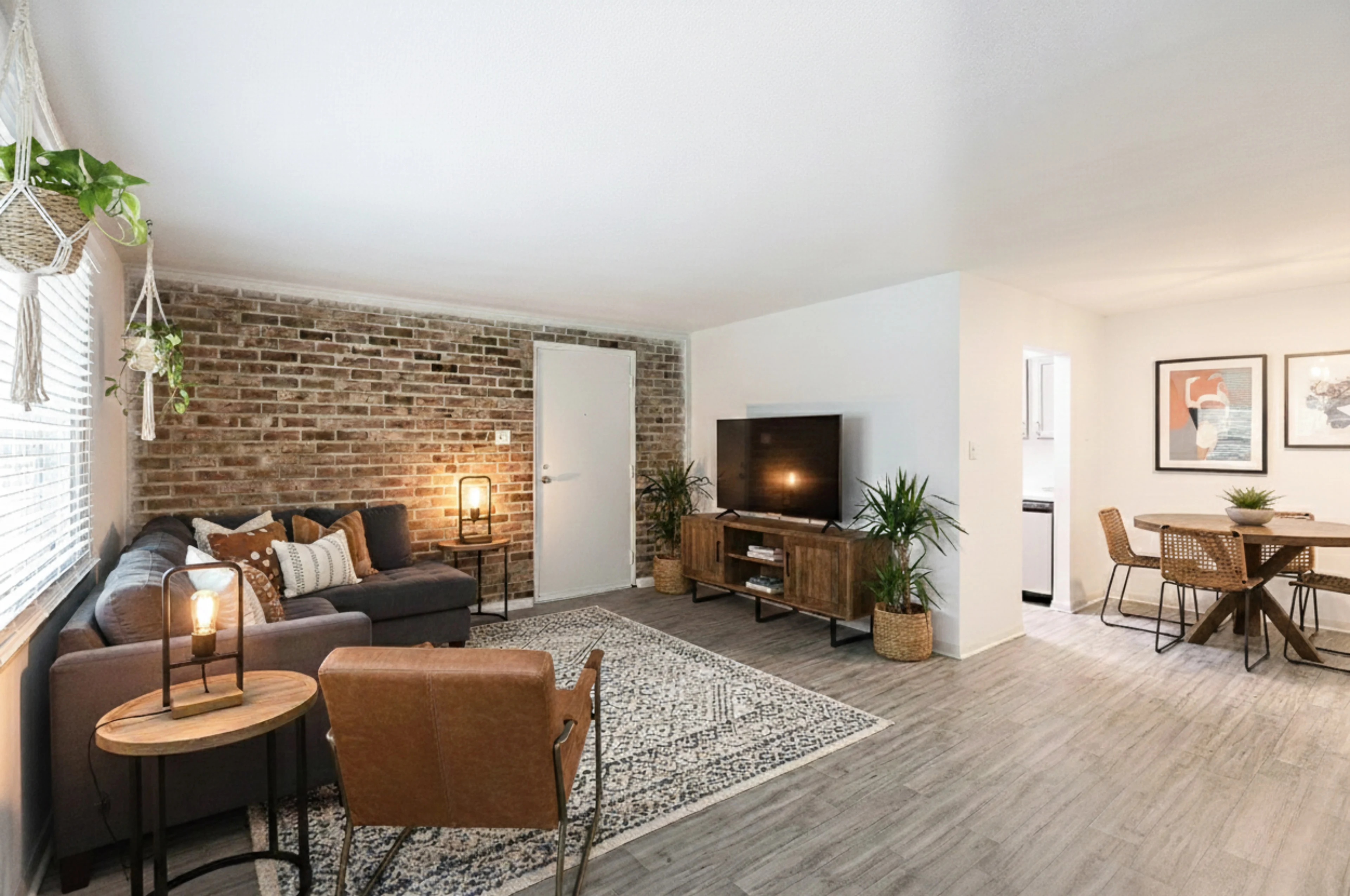 A cozy modern living room featuring a brick wall, a grey sofa with decorative cushions, a flat-screen TV, wooden furniture, and a dining area with a table and chairs.
