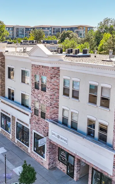 A street view of a row of commercial buildings with mixed architectural styles, showcasing brick and stucco facades, lined with trees and sidewalk, with parked cars along the curb.