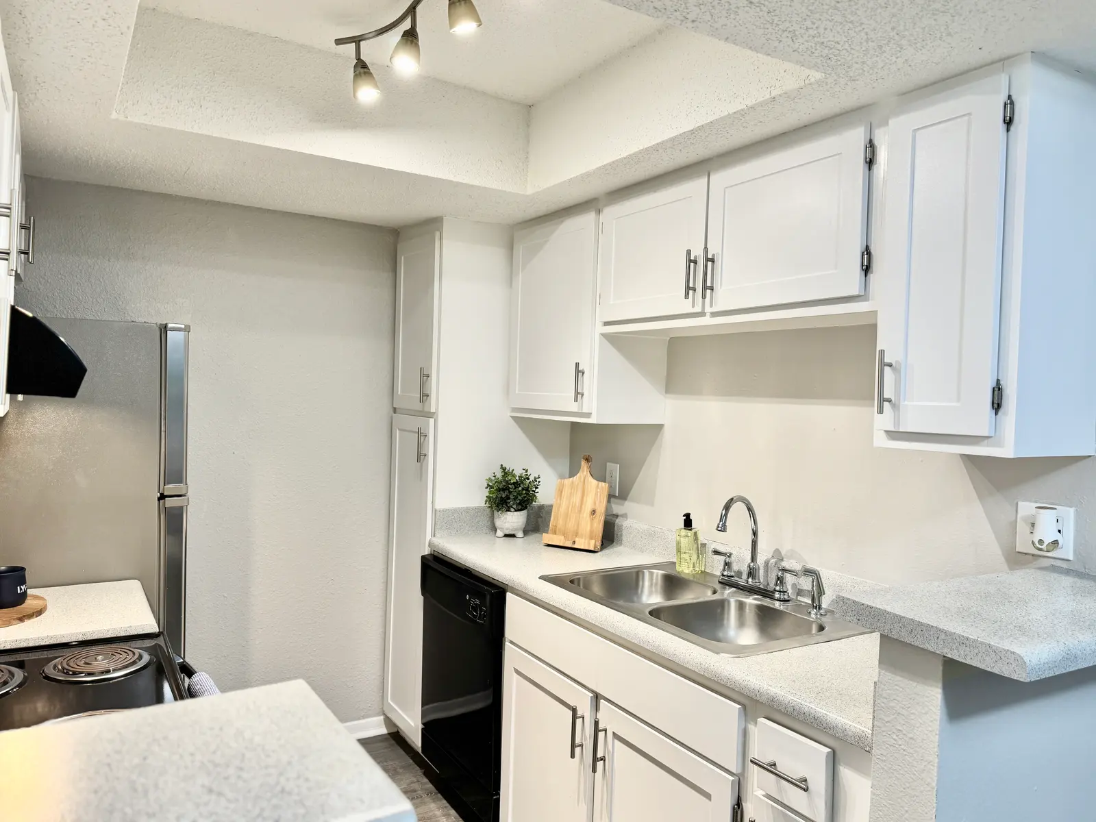 A modern kitchen featuring white cabinets, a double sink, and stainless steel appliances. The countertop is light-colored with a wooden cutting board and a small green plant. An overhead light fixture illuminates the space, and the flooring is dark.