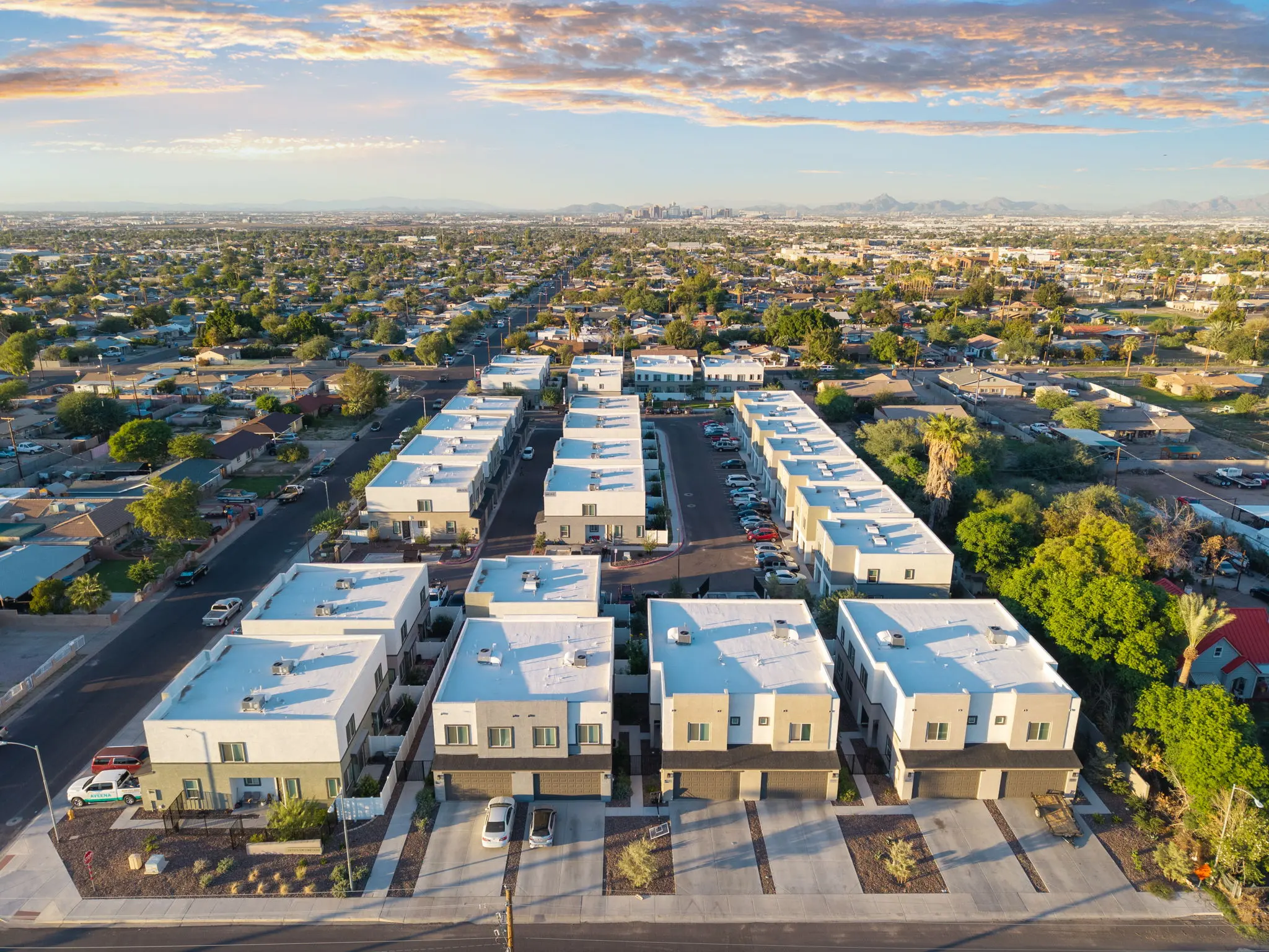 Aerial view of a modern housing development with multiple white-roofed buildings arranged in neat rows, surrounded by green trees and a suburban landscape.