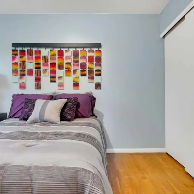 A modern bedroom featuring a bed with gray and purple bedding, a decorative wall hanging composed of colorful panels above the bed, and a bedside lamp. The walls are painted in soft blue, and a closet door is visible to the right.