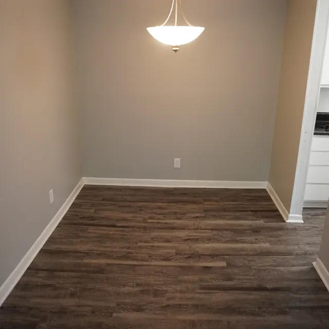 A spacious, empty room with light brown wooden flooring and gray walls. A modern pendant light hangs from the ceiling in the center of the room, creating a minimalist atmosphere. To the right, there is a glimpse of a kitchen area with white cabinetry.