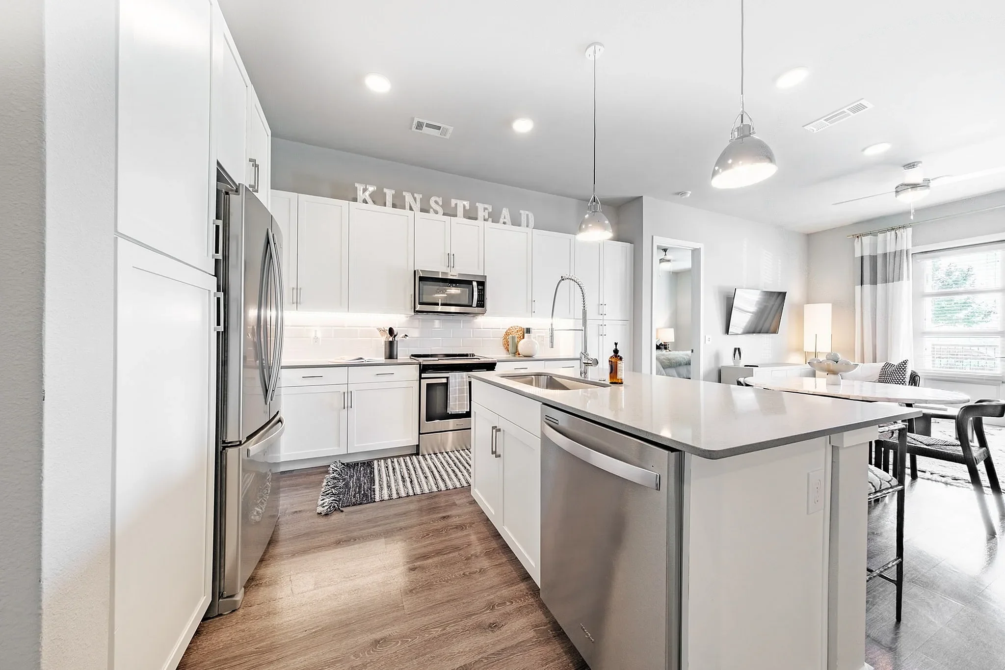 Modern Kitchen Interior A modern kitchen with white cabinetry, stainless steel appliances, and a spacious island. The room features bright lighting and a cozy atmosphere, with decorative elements like a rug and wall lettering.