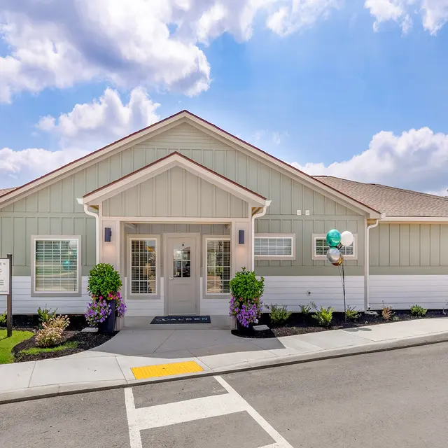 A modern one-story building with a light green exterior and white trim, featuring large windows and a welcoming entrance. The landscaping includes neatly trimmed shrubs and colorful flowers. Balloons in gold, teal, and white adorn the entrance, and the sky is partly cloudy.