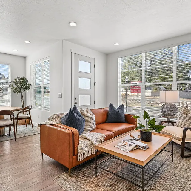 A cozy and modern living room featuring a brown leather sofa with decorative pillows, a beige armchair, a wooden coffee table with a plant, and a dining area with wooden chairs and a round table. Large windows provide natural light.